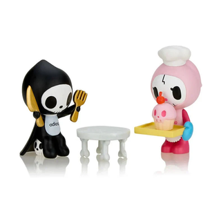 Bone Appetit figure design of Adios sitting in front of a table holding a golden fork and knife while Ciao Ciao is holding a tray with a skull-shaped cupcake on it while wearing a chef hat.