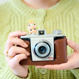 Close up image of a person holding a vintage camera with a small Sonny Angel Peacock figurine on top, person is wearing a green sweater.